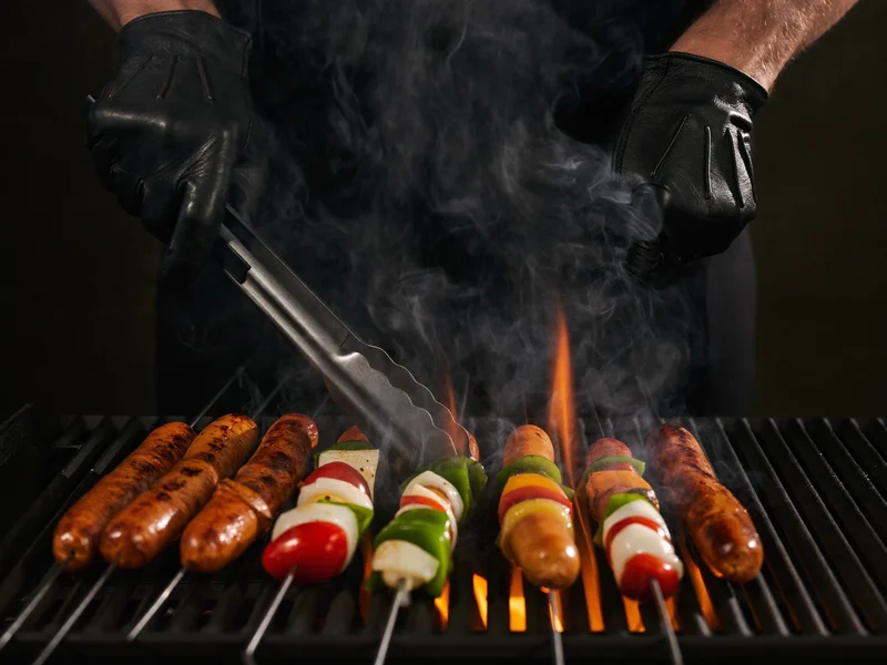 Illustration: Hände in Lederhandschuhen wenden Bratwürste und Gemüsespiesse über offenem Feuer